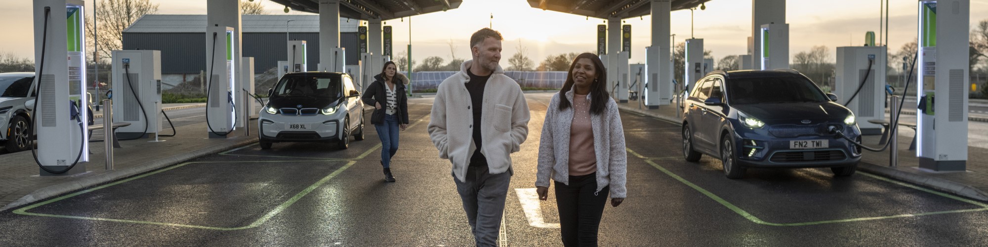 A couple walks across a forecort at an EV charging station.