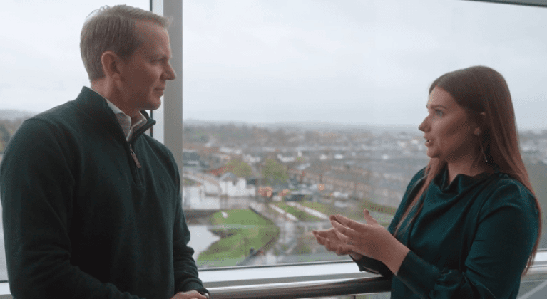 CEO Charlie Nunn and Cameron, an Economic Crime Analyst, stand in front of a window overlooking Belfast. They are discussing her experience stopping fraudsters.